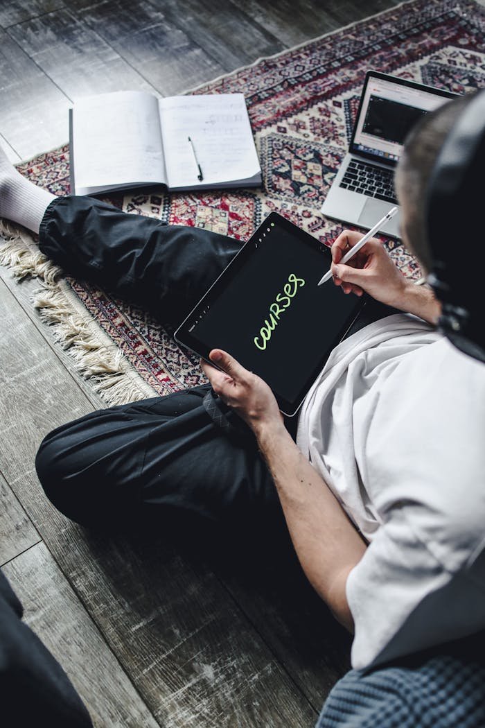 A young adult using a tablet to write 'courses' while seated on a patterned rug indoors.